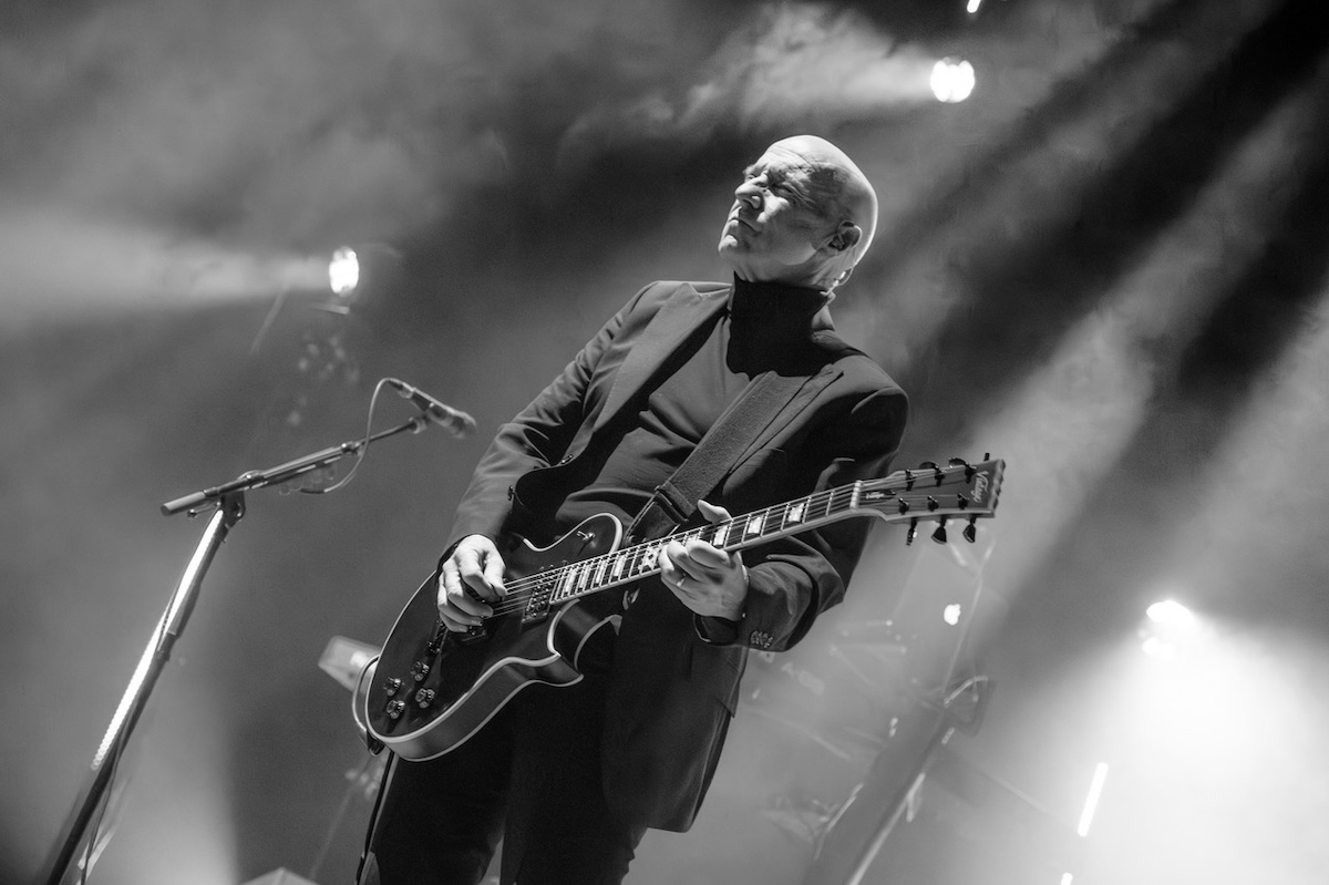 Black and white live performance photo of musician Midge Ure playing electric guitar under bright stage spotlights.